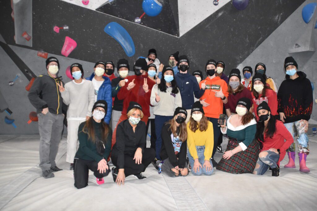 Group Photo of Boulderz Staff Climbing wall behind them, 27 people back row standing, front row kneeling down. All wearing face masks and black hats with "Boulderz"