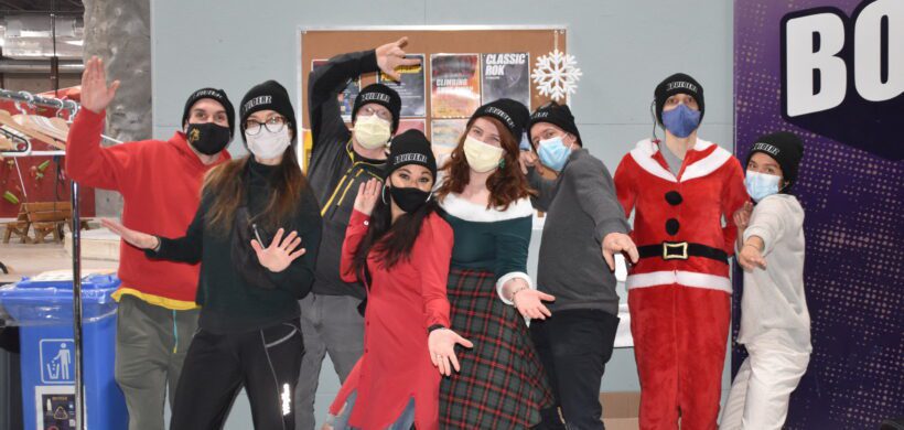 Seven People n face masks and hats that have the text "Boulderz" on them standing in a line side by side making funny hand gestures.